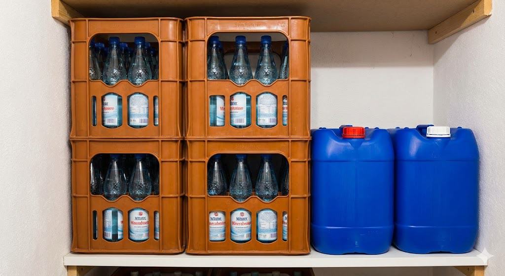 Ein Regal im Keller mit gestapelten Wasserkästen und zwei blauen Trinkwasserkanistern als Notvorrat.